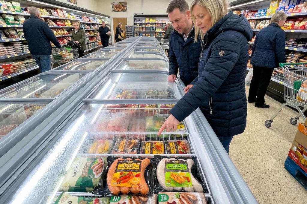 Shoppers choosing Sausage Haus pork bratwurst and cheese frankfurter in a Farmfoods frozen food aisle