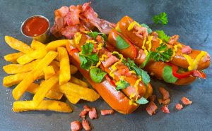 Bacon Frankfurter hotdogs topped with mustard, crispy bacon, and parsley, served with golden fries and ketchup — showcasing premium German sausages wholesale UK.