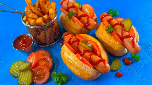 Three pork hotdogs in buns with ketchup, pickles and tomato, served with chips on a blue background