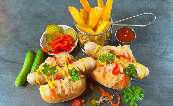 Two German Bratwurst hot dogs with sauerkraut, mustard, and pickles served with fries and ketchup on a slate background.