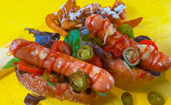 German sausage hotdog topped with melted cheese, jalapeños, ketchup, and fresh greens on a yellow background.