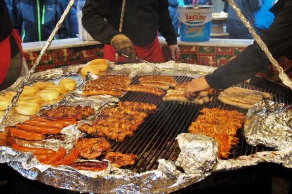 Chefs grilling German sausages, skewers, and buns at a busy Christmas market BBQ stall, showcasing festive catering with authentic German flavours.