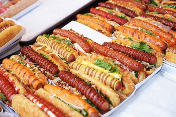 Rows of grilled German sausages in hotdog buns with colourful sauces and toppings, prepared for catering at a busy BBQ or event.