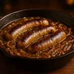 Bratwurst in beer and onion gravy served in a rustic cast-iron skillet with caramelised onions and fresh herbs