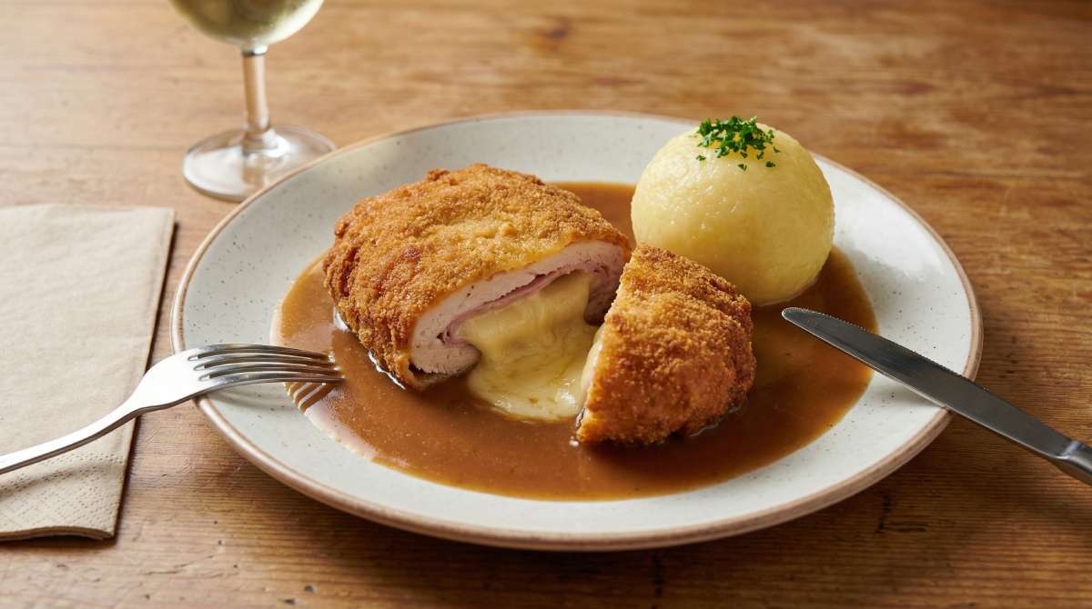 German-style cordon bleu cut open with melted cheese and ham, served in brown gravy with a potato dumpling on a speckled plate