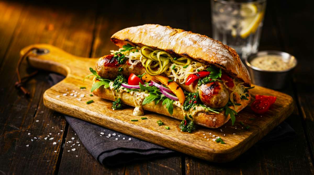 Elevated hotdog sandwich with browned Bratwurst in a crusty artisan roll, topped with rocket, sauerkraut, peppers, pickles and creamy sauce on a wooden board.