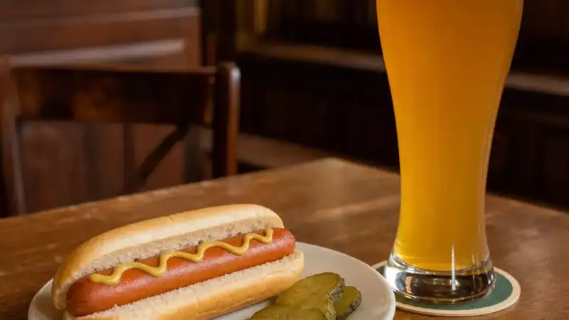 A mild frankfurter with mustard and pickles is paired with a wheat beer in a pub setting.