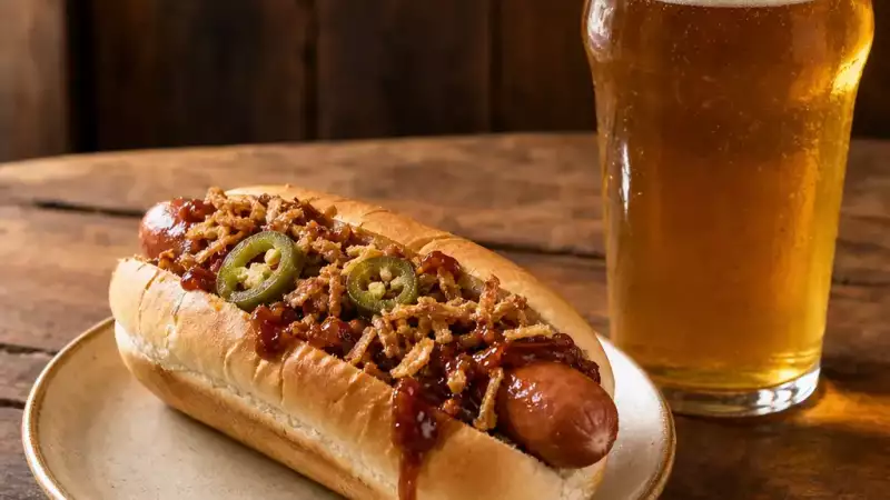 A chilli frankfurter with jalapeños and crispy onions sits beside a pint of pale ale on a pub table.