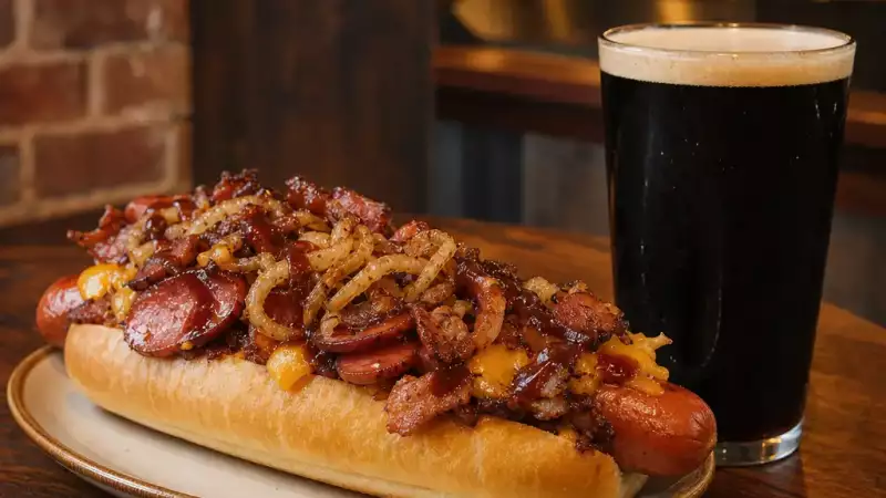 A loaded hot dog with smoky toppings served beside a dark pint, showing a hearty beer pairing for a pub menu.