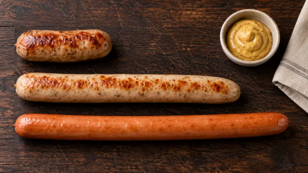A short British-style banger beside a longer pale bratwurst and smooth frankfurter on a dark wooden board.